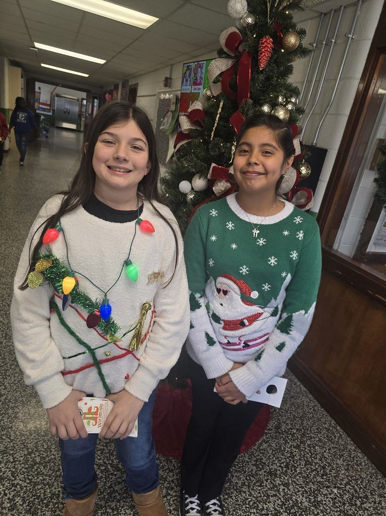 Two elementary students stand together smiling in a school hallway beside a decorated Christmas tree, wearing festive holiday sweaters.