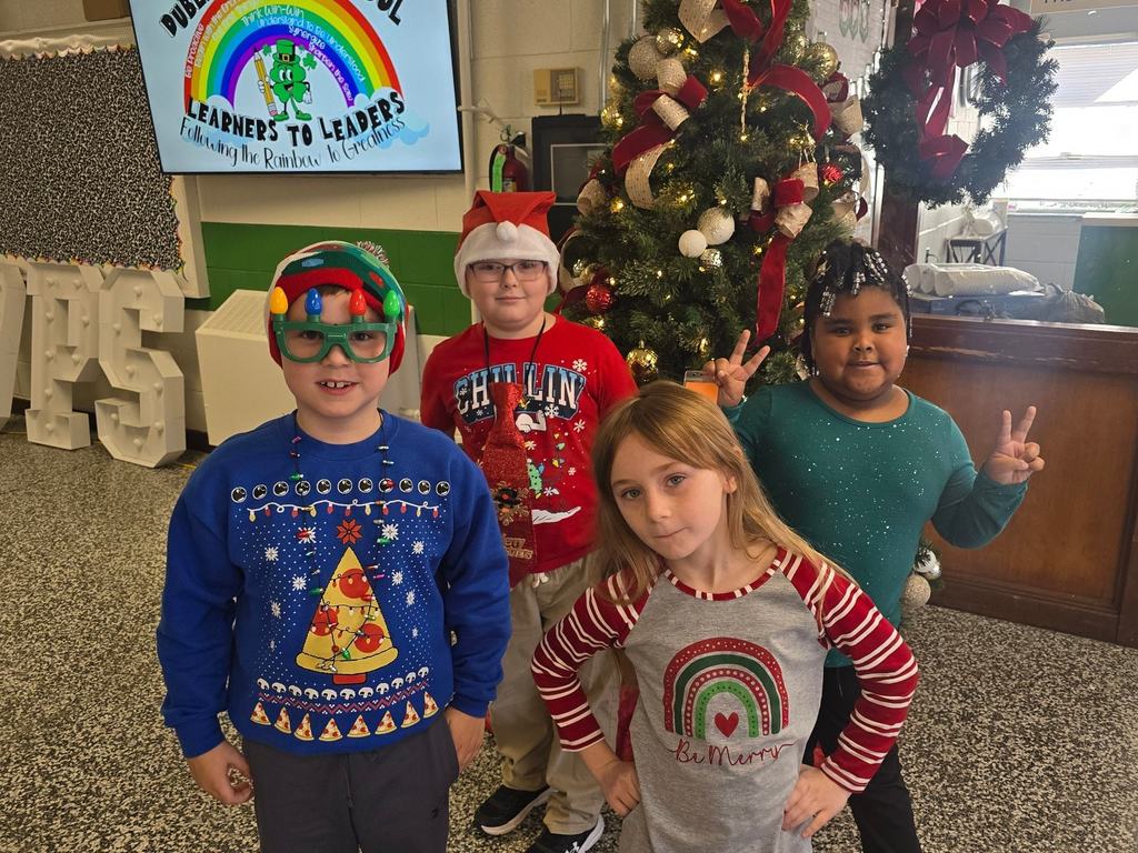 Five elementary students pose together in front of a Christmas tree, wearing colorful holiday sweaters and accessories inside a school lobby.