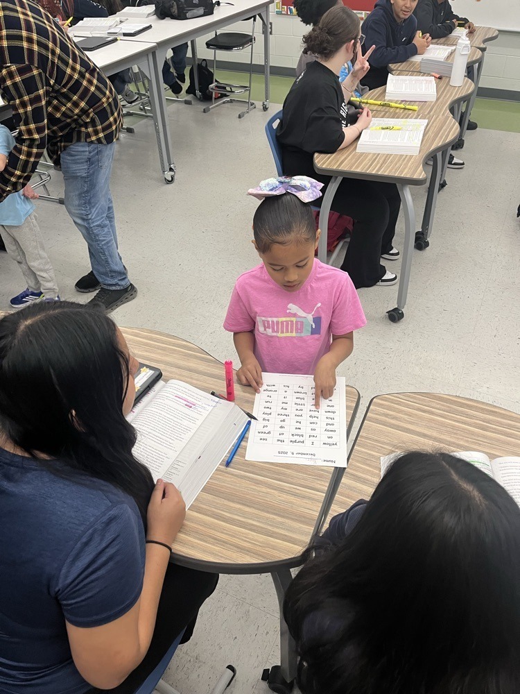 A young student leads a small-group reading activity while classmates follow along using worksheets at their desks.