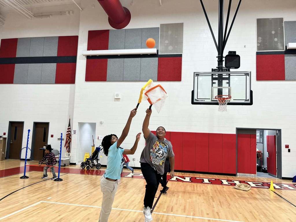 Students use handheld nets to catch a ball during a cooperative physical education activity in the gym.