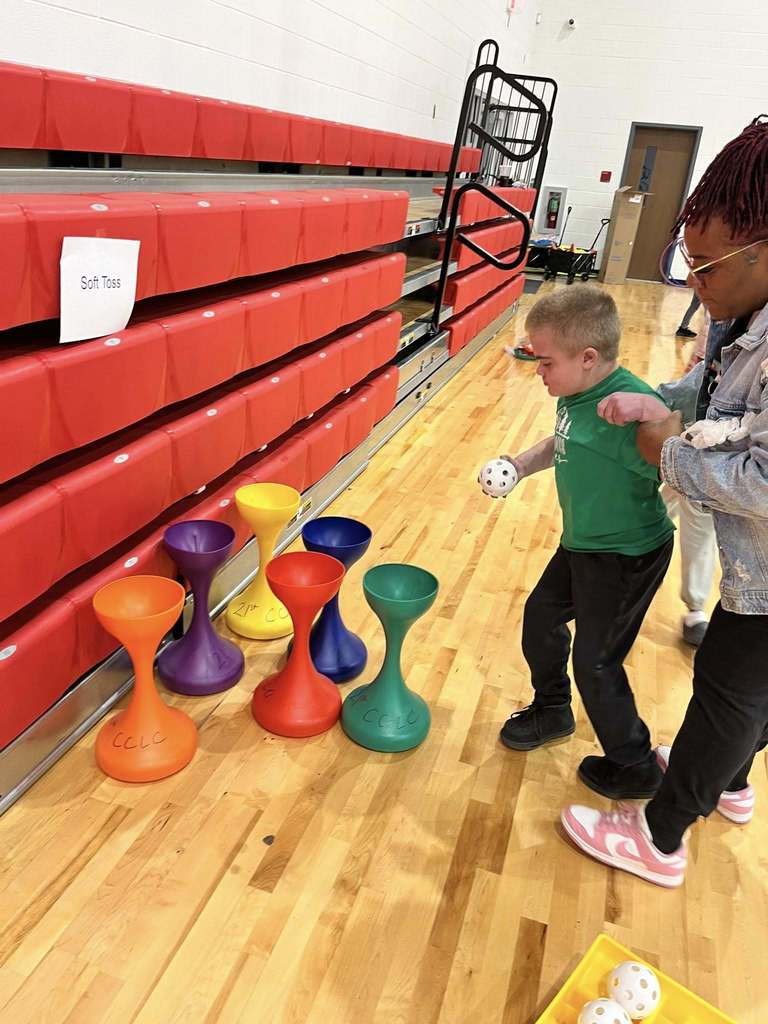 Student participates in a soft-toss activity during an adaptive physical education station in the school gym.