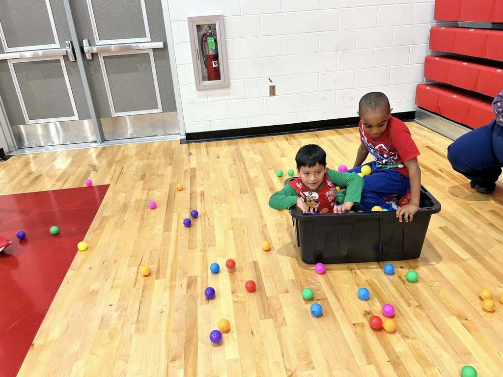 Two students sit inside a bin filled with colorful balls during a playful field day sensory activity in the gym.