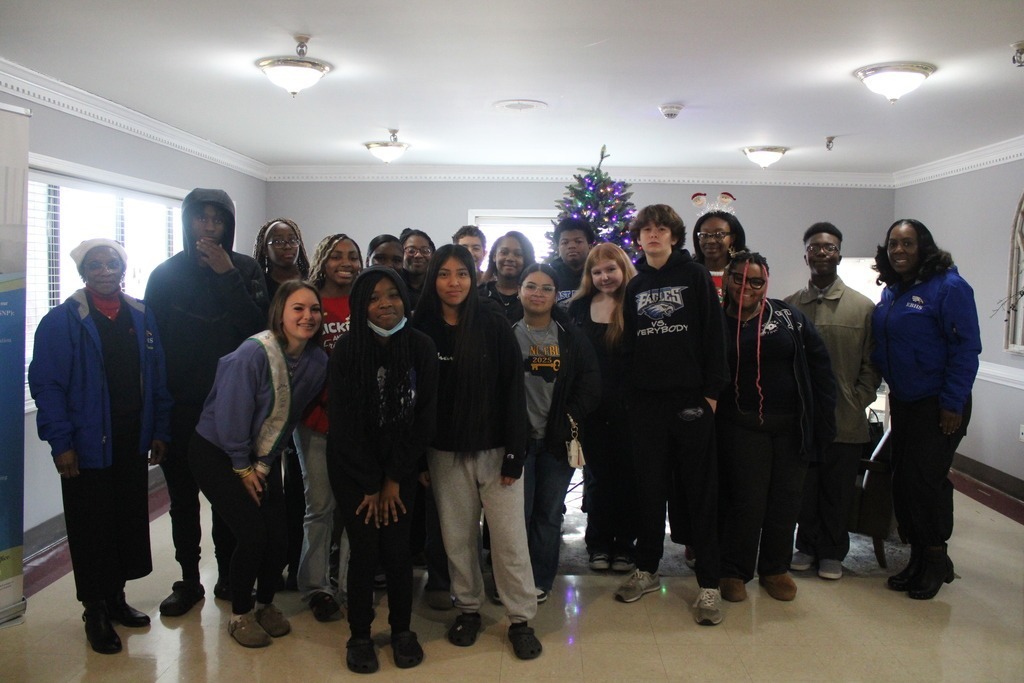 A group of high school students stand in a hallway holding gift bags, smiling as they prepare to participate in a holiday service activity.