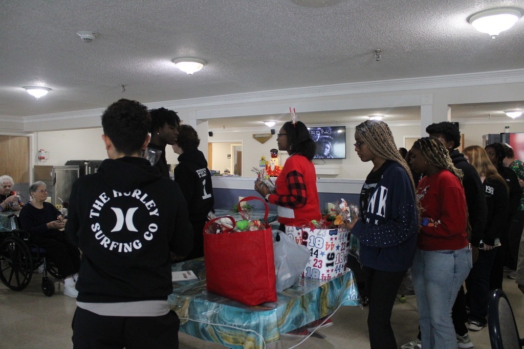 Students gather in a common room decorated for the holidays, standing near tables with gift bags and craft supplies during a community outreach event.
