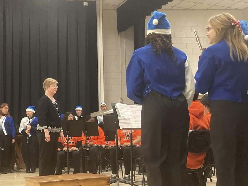 A band director stands on a podium conducting a student band during a winter concert, while musicians in blue-and-white uniforms prepare to play on stage.