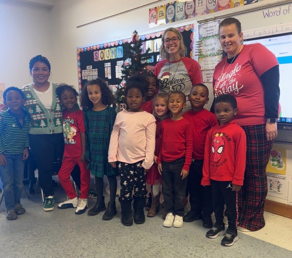 A group of students and teachers pose in a classroom decorated for the holidays, standing near a Christmas tree and classroom bulletin boards.