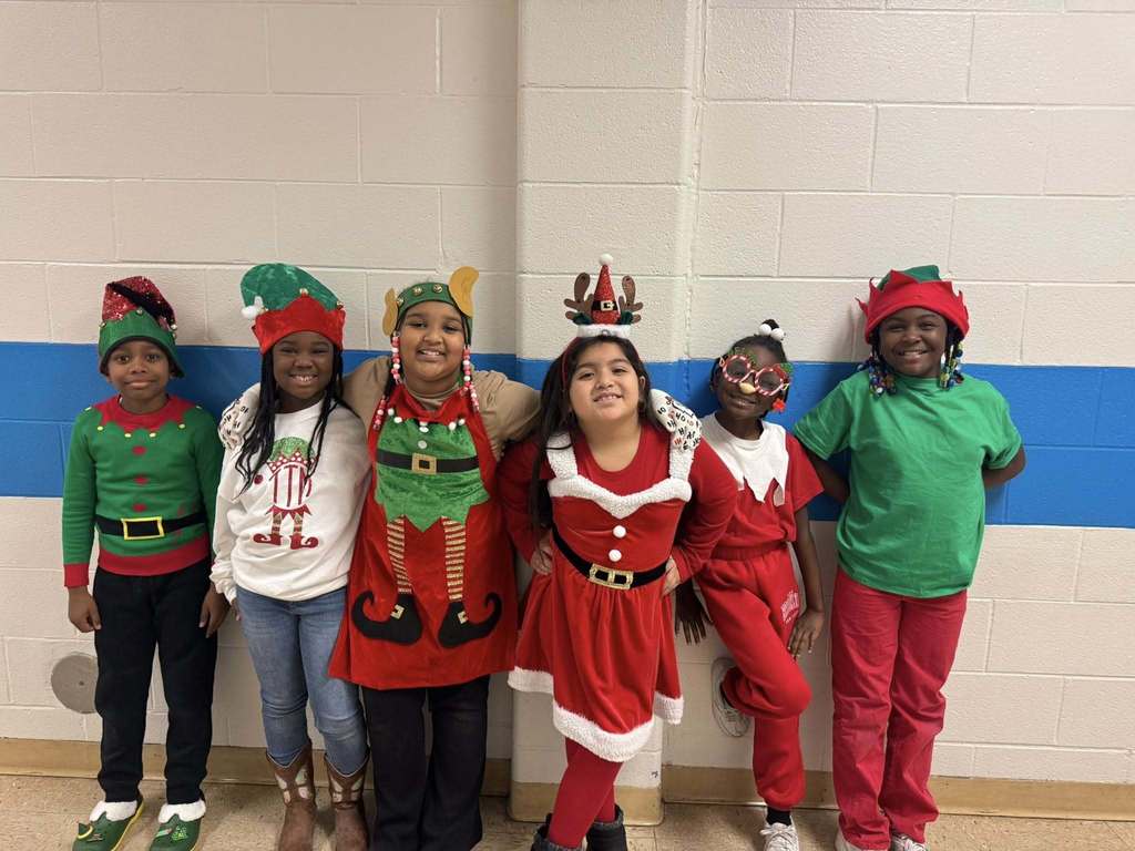 A group of elementary students stand in a hallway wearing festive holiday outfits and elf hats, smiling and posing together against a blue-and-white wall.