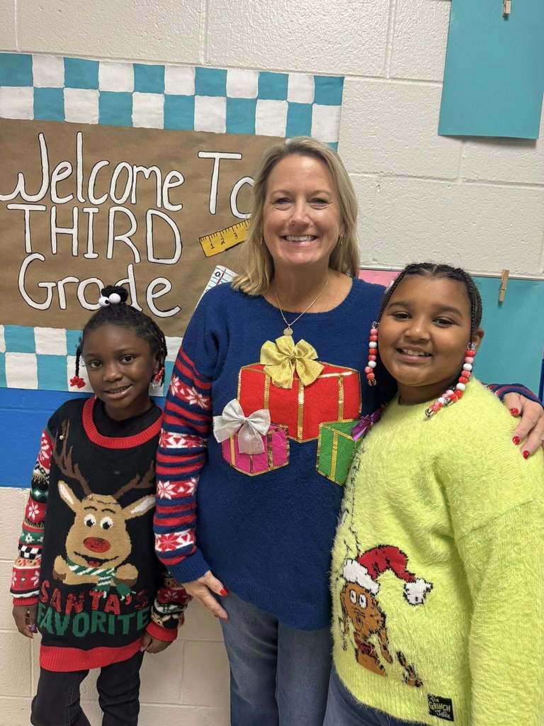 A teacher stands between two students in a school hallway, all smiling and wearing colorful holiday sweaters in front of a “Welcome to Third Grade” sign.