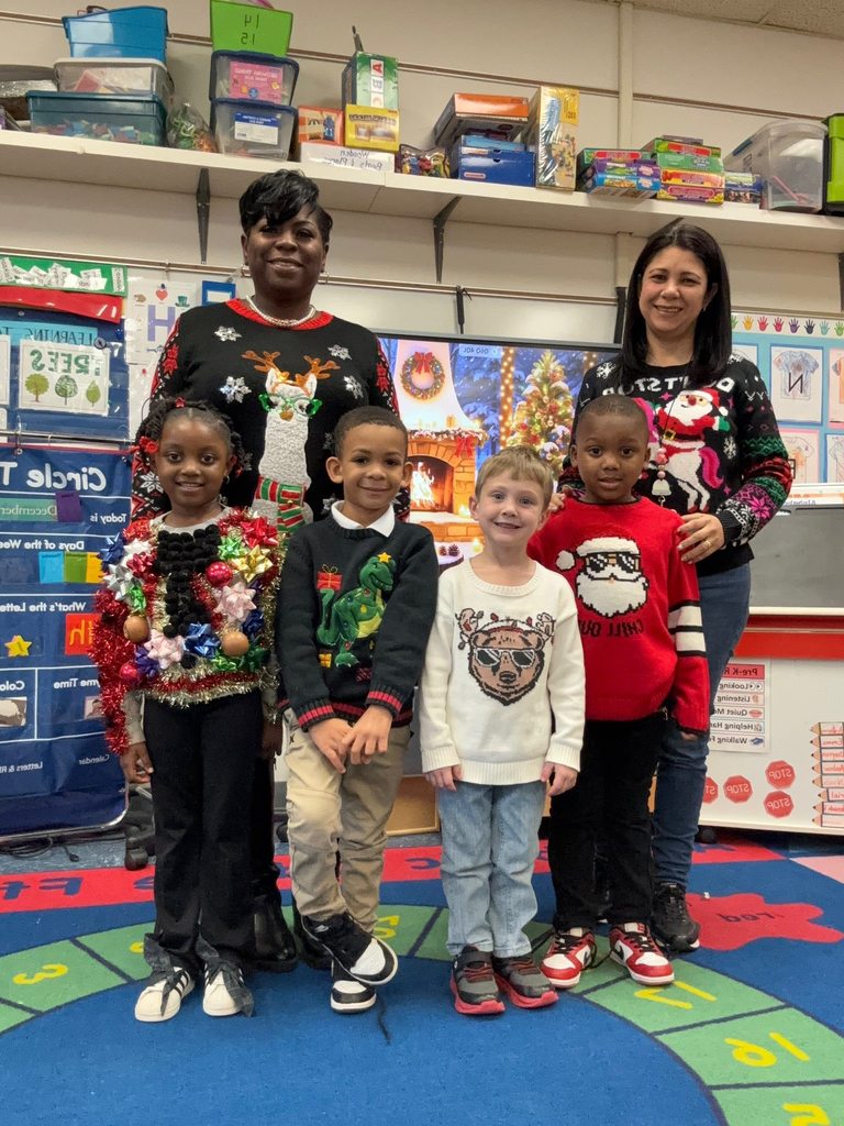 Two teachers stand behind a group of young students in a classroom, all wearing holiday-themed sweaters and smiling for a festive group photo.