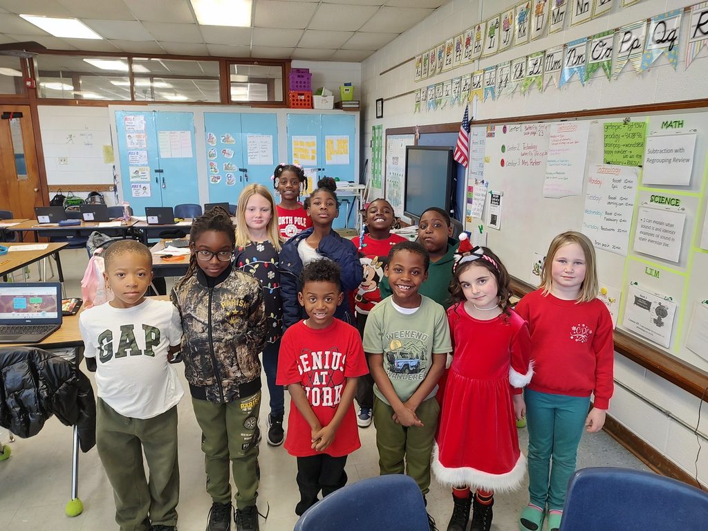A class of elementary students stand together in a classroom wearing red and green holiday clothing, smiling and facing the camera.
