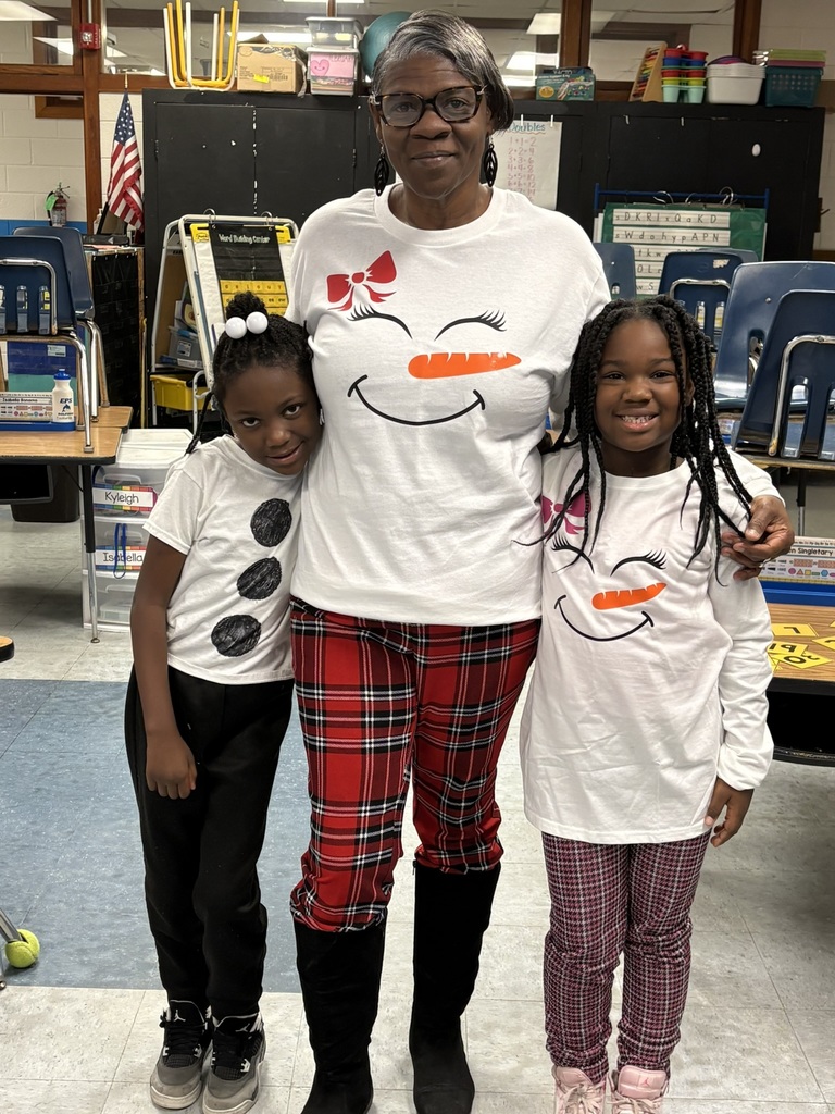 A teacher stands with two students in a classroom, all wearing matching snowman-themed shirts and smiling for a holiday photo.