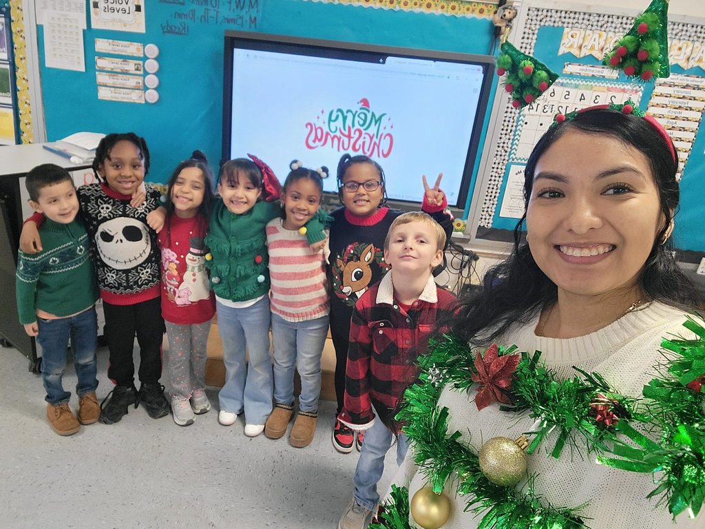 A teacher takes a classroom selfie with students wearing holiday sweaters and accessories, standing in front of a screen that reads “Merry Christmas.”