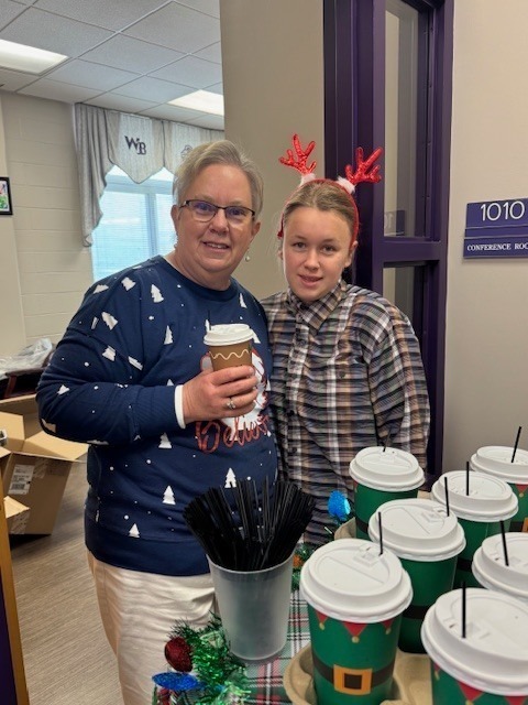 A teacher and student stand behind a holiday coffee station with cups and stirrers, both smiling and wearing festive attire and reindeer antlers.