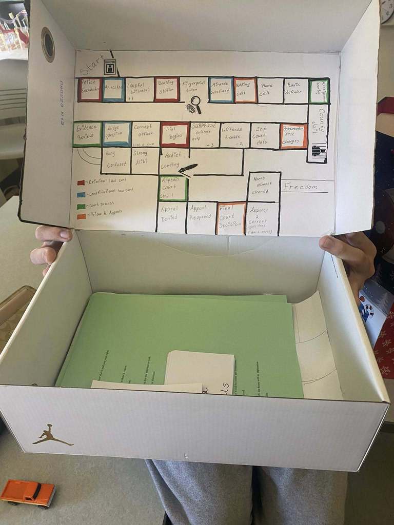 Handmade board game displayed inside a shoebox, illustrating the criminal justice process with labeled steps, color-coded sections, and game pieces for a civics class project.