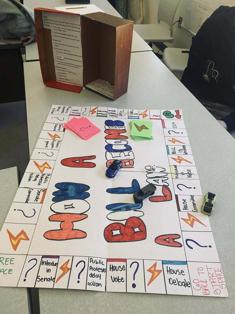 Student-created board game titled “How a Bill Becomes a Law,” laid out on a classroom table with dice, toy cars as game pieces, and labeled spaces showing steps in the legislative process.