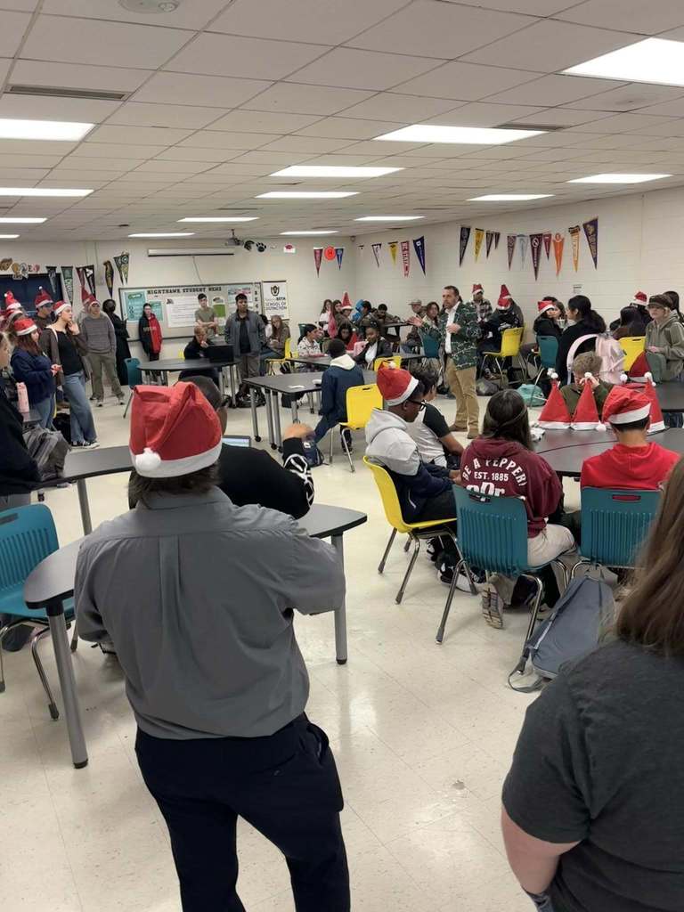 Students wearing Santa hats sit and stand around tables as an instructor addresses the room during a holiday classroom activity.