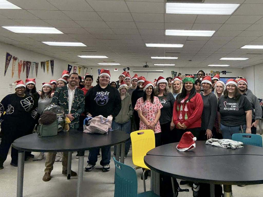 A large group of students and staff wearing Santa hats pose together in a classroom decorated with college pennants.