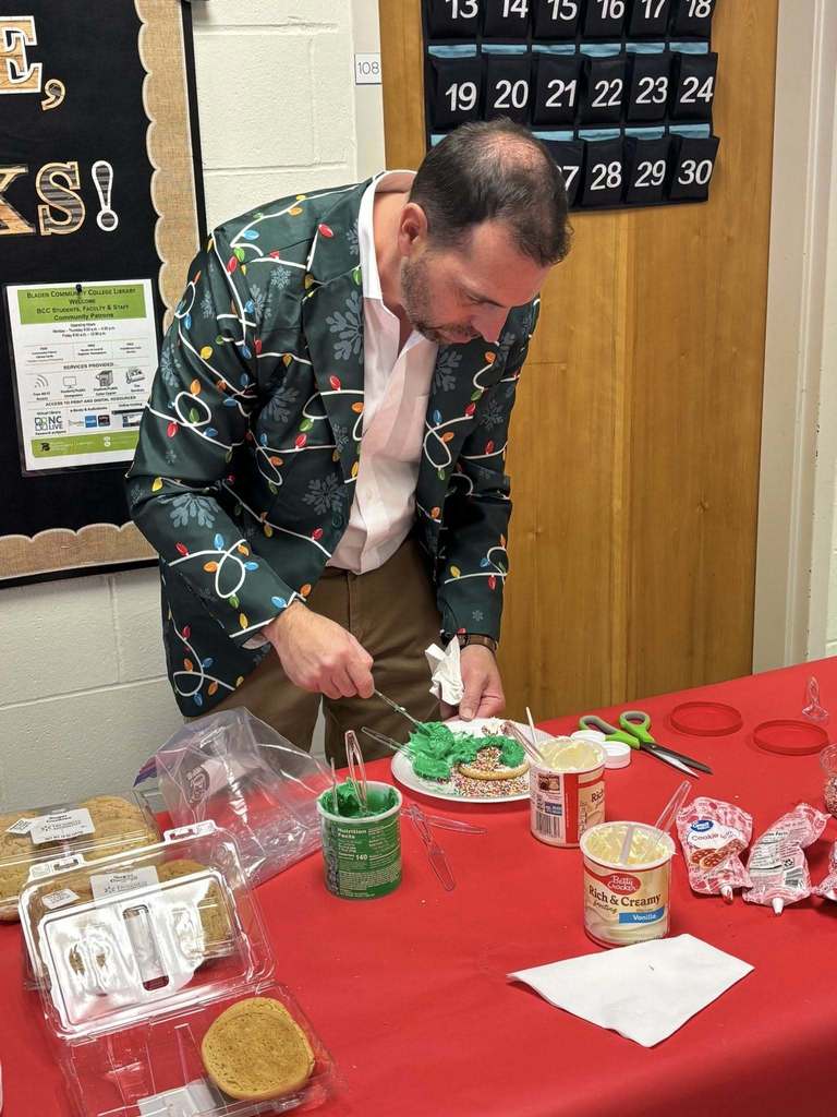 An instructor wearing a festive jacket decorates sugar cookies with green icing at a holiday-themed table covered with baking supplies.