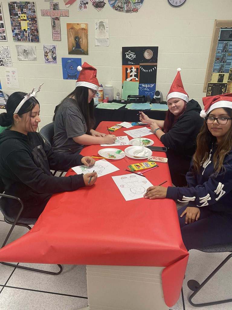 Four students wearing Santa hats sit at a red-covered table coloring holiday-themed worksheets with crayons and markers.