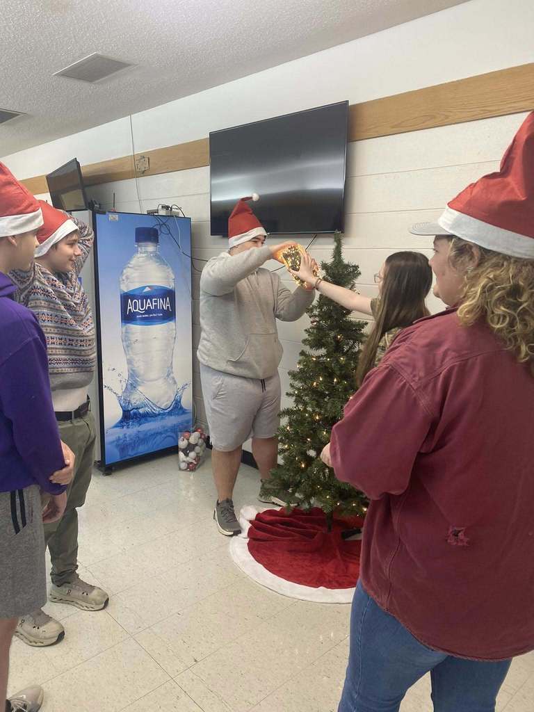 Students wearing Santa hats gather around a small Christmas tree as one student places a lighted ornament on it.
