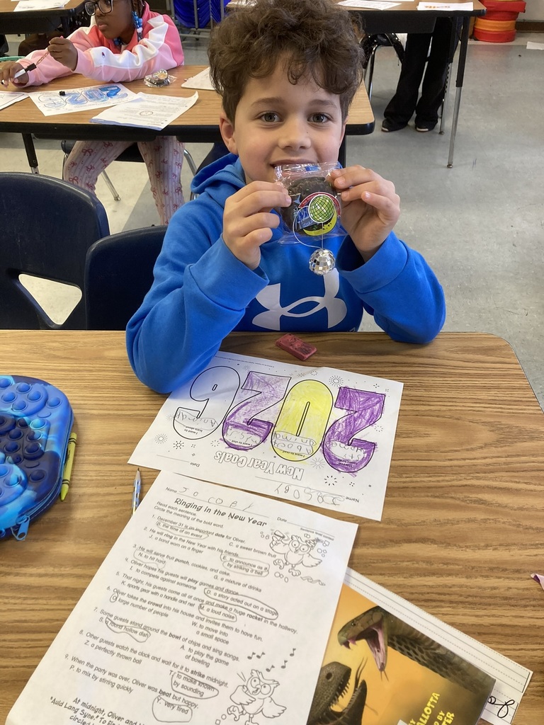 A student smiles while holding up a New Year’s themed treat in front of their “2025” goals worksheet. Additional classwork and supplies are visible on the desk, showing a goal-setting activity in progress.