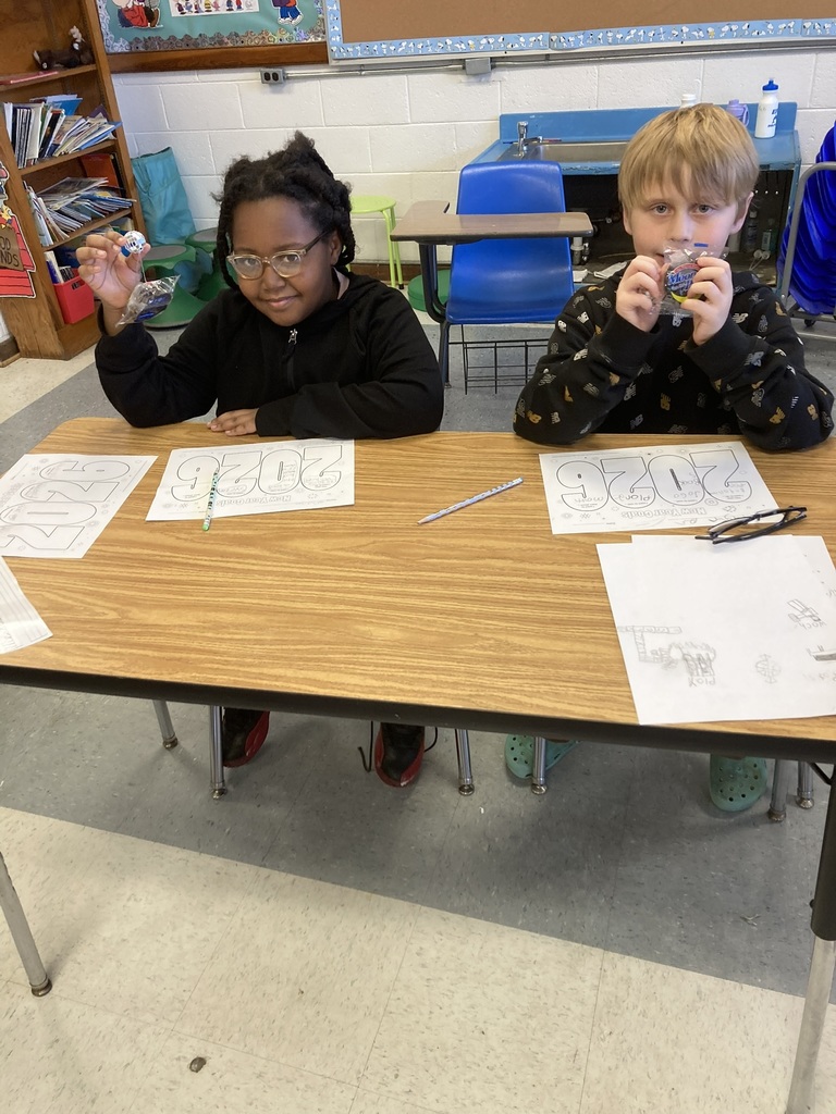 Two students sit at a classroom table holding up small New Year’s themed treats while smiling. Each student has a “2025” goals worksheet in front of them on the desk.