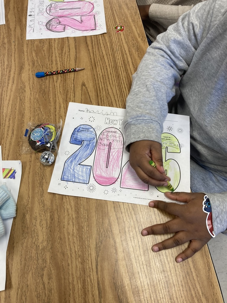 A student sits at a classroom table coloring a “2025” New Year goals worksheet with a green crayon. The student’s paper is brightly colored, and a small New Year’s themed treat is on the table beside the worksheet.