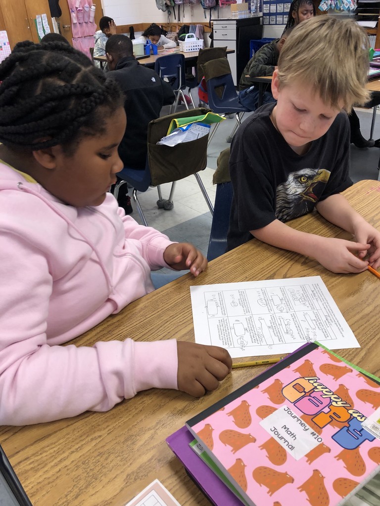 Two students sit side by side at a classroom table reviewing a math worksheet. One student looks at the paper while the other listens, demonstrating cooperative learning.