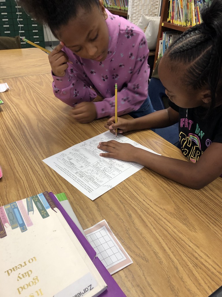 Two students sit at a table working together on a math worksheet. One student writes while the other leans in, pointing and offering support as they discuss the problem.