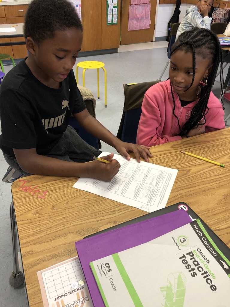 Two students work together at a desk during math practice. One student writes on a worksheet while the partner watches closely, ready to coach and assist.