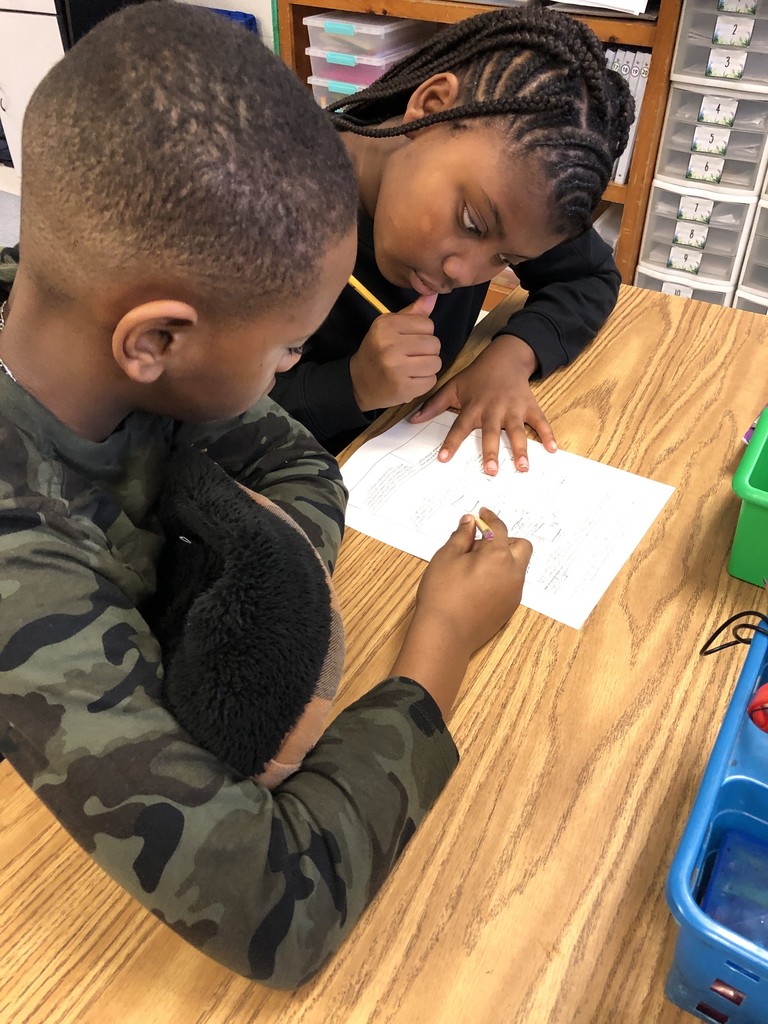 Two students sit closely at a table focused on a shared math worksheet. One student points to the paper while the other follows along, showing teamwork and problem-solving.