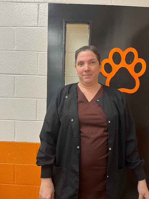 A staff member stands in a school hallway in front of a black door with an orange cougar paw, wearing a dark jacket and smiling at the camera.
