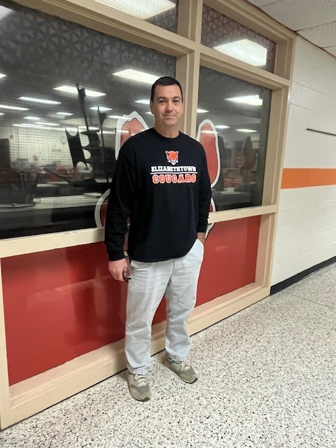 A staff member wearing an Elizabethtown Middle School Cougar sweatshirt stands in a school hallway in front of a glass display, smiling and posing for a photo