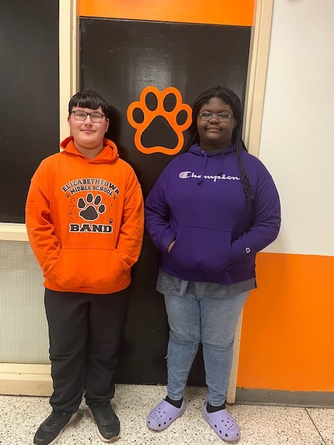 wo students stand side by side in a school hallway in front of a black door with an orange cougar paw, smiling and posing for a photo.
