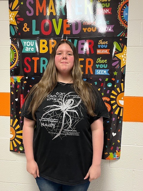 A student stands in a school hallway in front of a colorful motivational poster, wearing a light green hoodie and smiling for the photo.