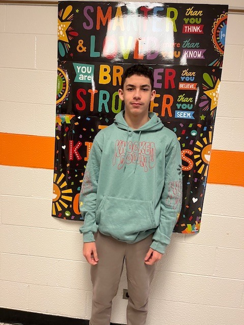 A student stands in a school hallway in front of a colorful motivational poster, wearing a light green hoodie and smiling for the photo.