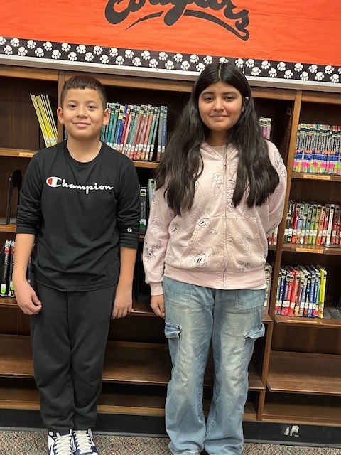Two students stand side by side in a school library in front of bookshelves filled with books, smiling and posing for a photo.