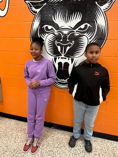 Two students stand in a school hallway in front of an orange wall featuring a black cougar mural, smiling and posing for a photo.