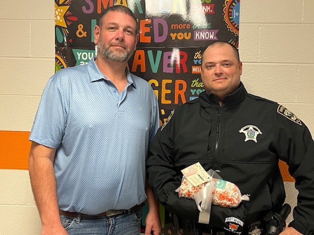 A school staff member stands beside a school resource officer in uniform in a school hallway, posing for a photo. The officer is holding a small gift as both smile at the camera.
