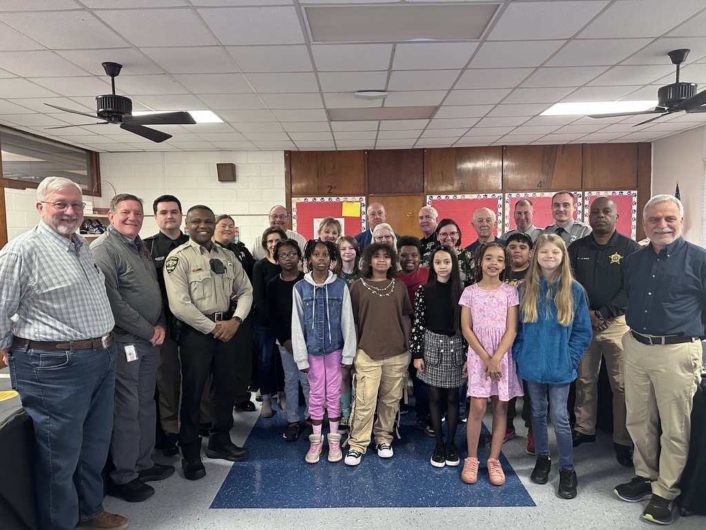 A group photo of elementary students standing in the front row with local law enforcement officers, school staff, and community members behind them inside a classroom, smiling for the camera during a school visit or recognition event.