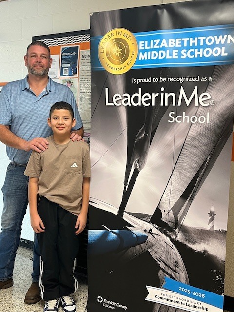 A school staff member stands beside a student in a school hallway in front of an Elizabethtown Middle School “Leader in Me” banner, posing together for a photo.