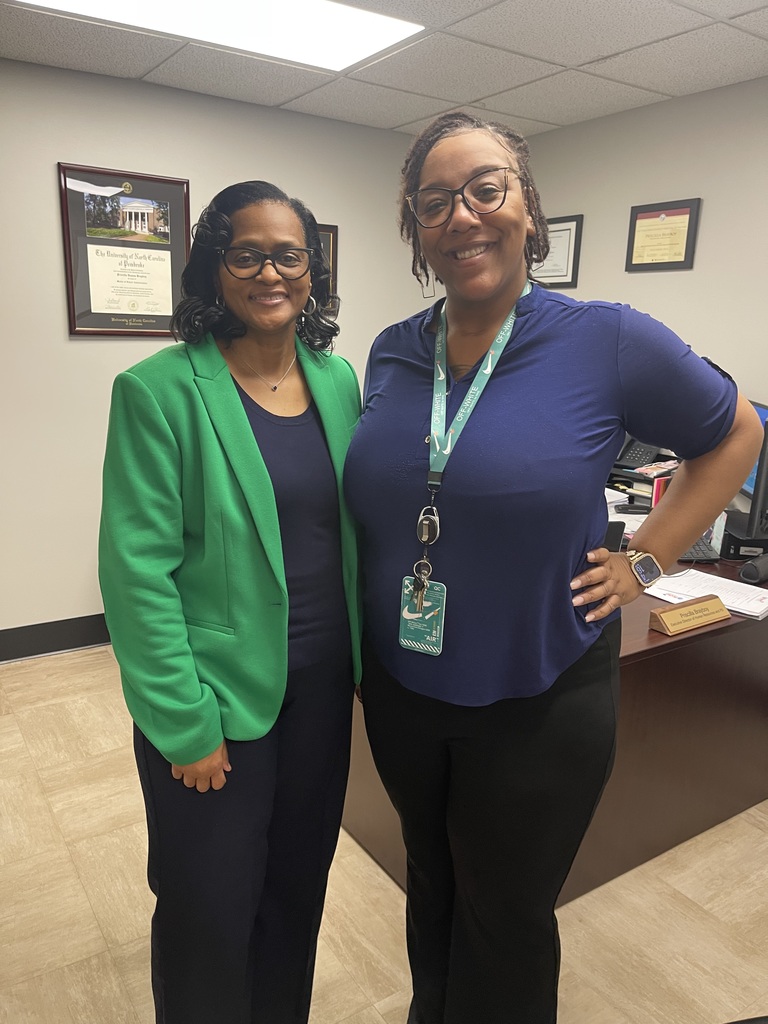 Two Bladen County Schools staff members smile while standing in an office, with framed certificates visible on the wall behind them.