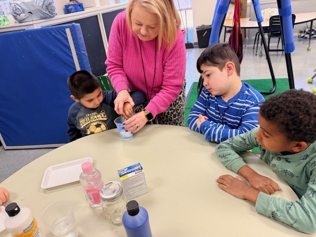 A teacher helps young students create a “snowstorm in a jar” at a classroom table, stirring a blue liquid in a clear container while the children watch and participate in the hands-on science activity.