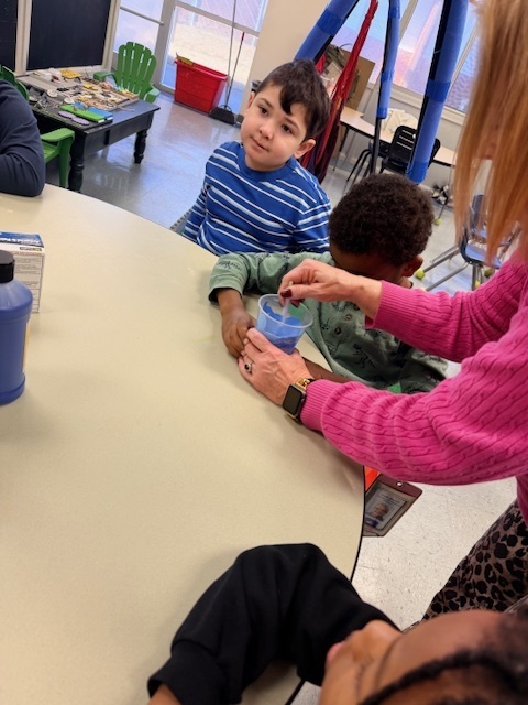 A teacher helps young students create a “snowstorm in a jar” at a classroom table, stirring a blue liquid in a clear container while the children watch and participate in the hands-on science activity.