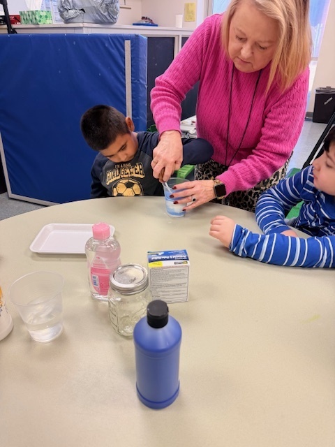 A teacher helps young students create a “snowstorm in a jar” at a classroom table, stirring a blue liquid in a clear container while the children watch and participate in the hands-on science activity.