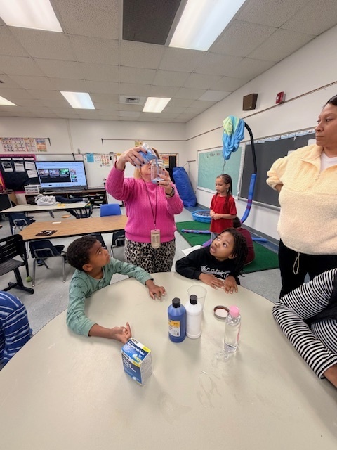 A teacher helps young students create a “snowstorm in a jar” at a classroom table, stirring a blue liquid in a clear container while the children watch and participate in the hands-on science activity.