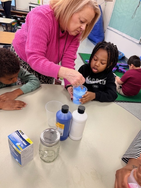 A teacher helps young students create a “snowstorm in a jar” at a classroom table, stirring a blue liquid in a clear container while the children watch and participate in the hands-on science activity.