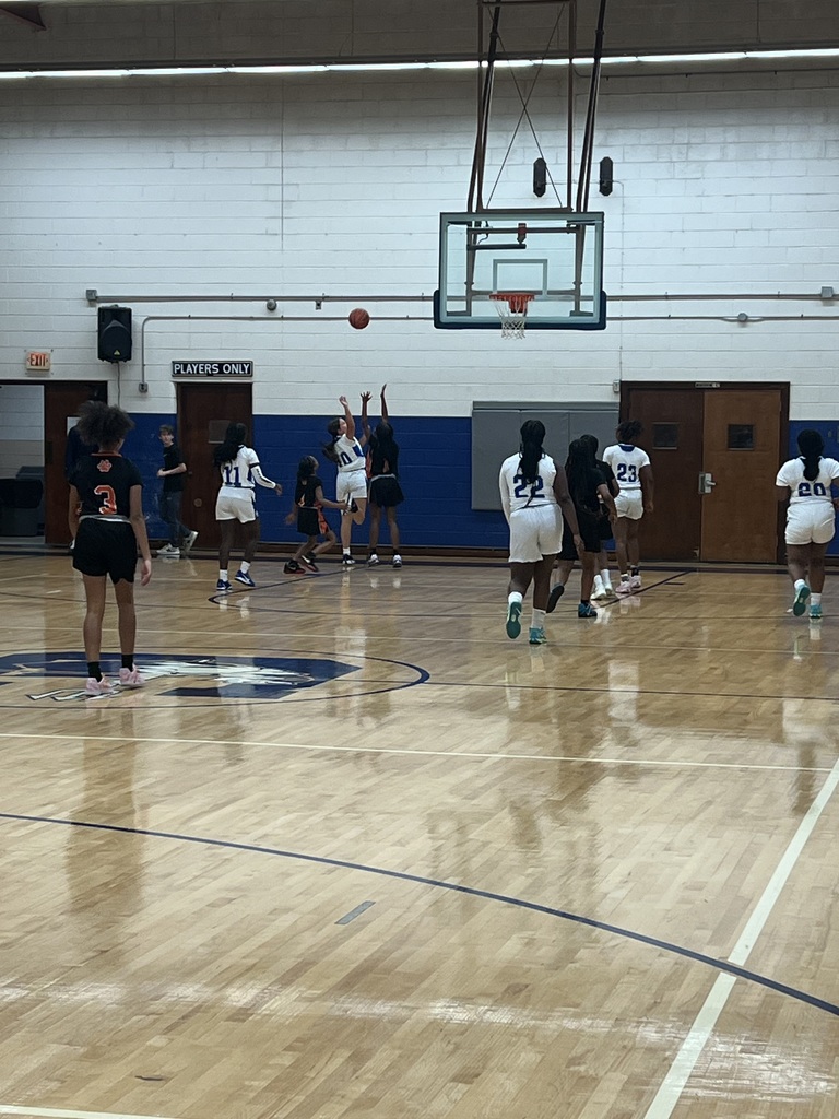 Girls basketball players move down the court as a shot arcs toward the basket during a fast-paced sequence of play.