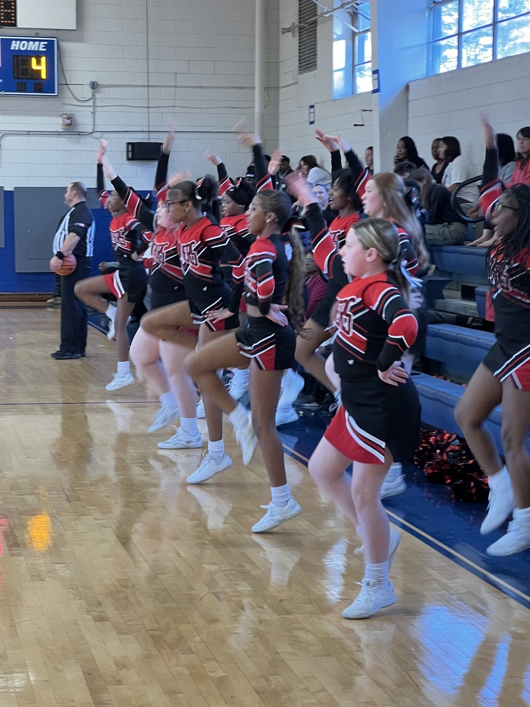 A cheerleading squad performs a synchronized routine on the sideline, raising their arms in unison during a basketball game
