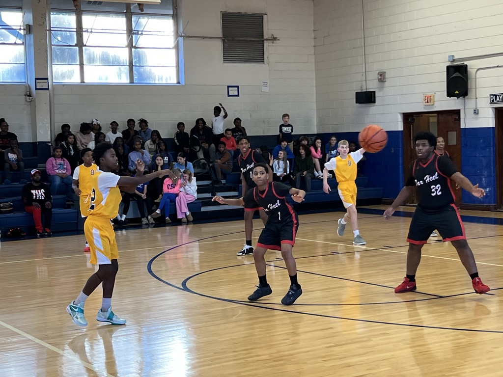 A boys basketball player passes the ball across the court while defenders react, with students seated in the bleachers behind the play.